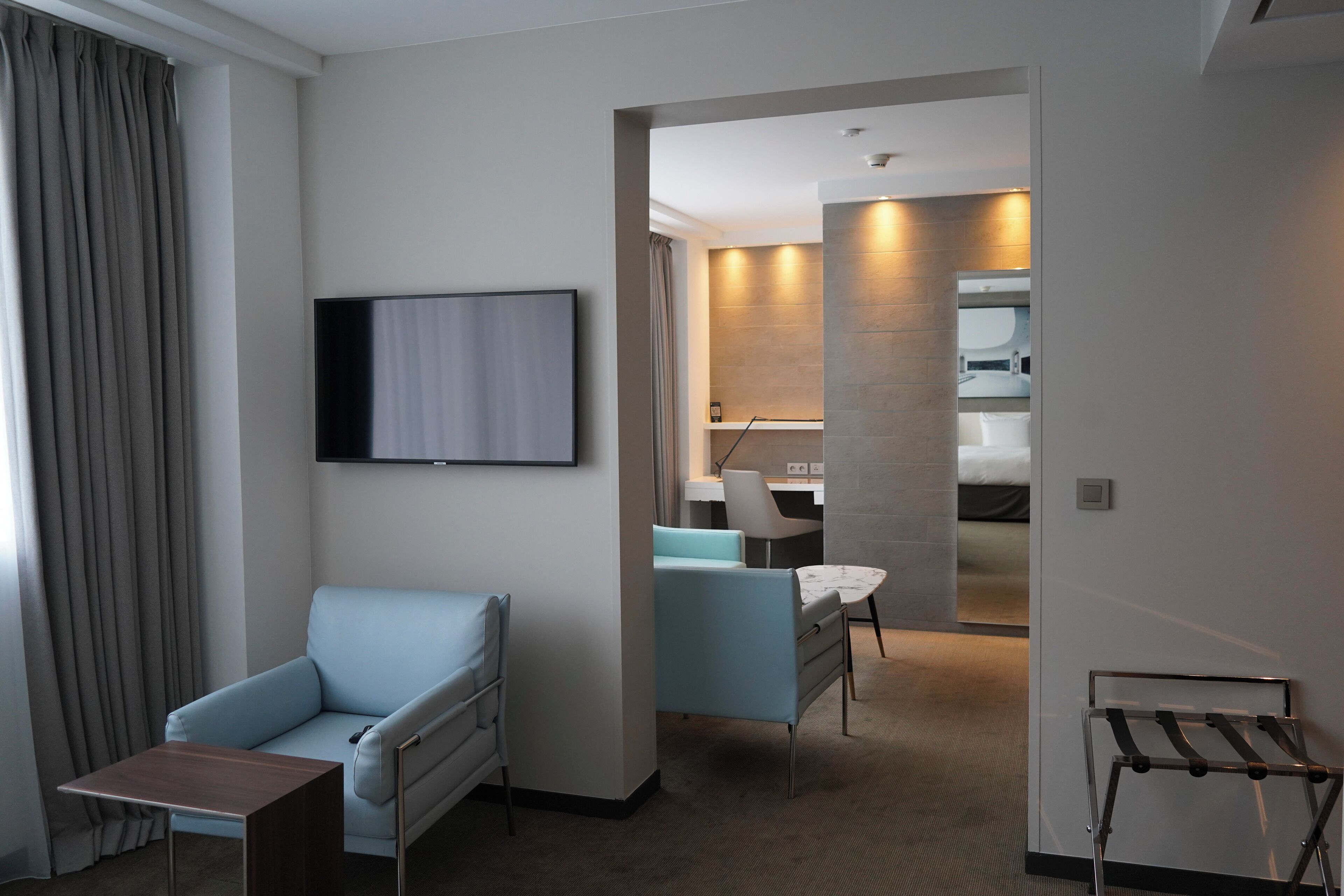 Una habitación de hotel moderna con sofá, una mesita, televisor montado en la pared, escritorio con silla y baño visible a través de una puerta abierta.