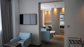 Una habitación de hotel moderna con sofá, una mesita, televisor montado en la pared, escritorio con silla y baño visible a través de una puerta abierta.