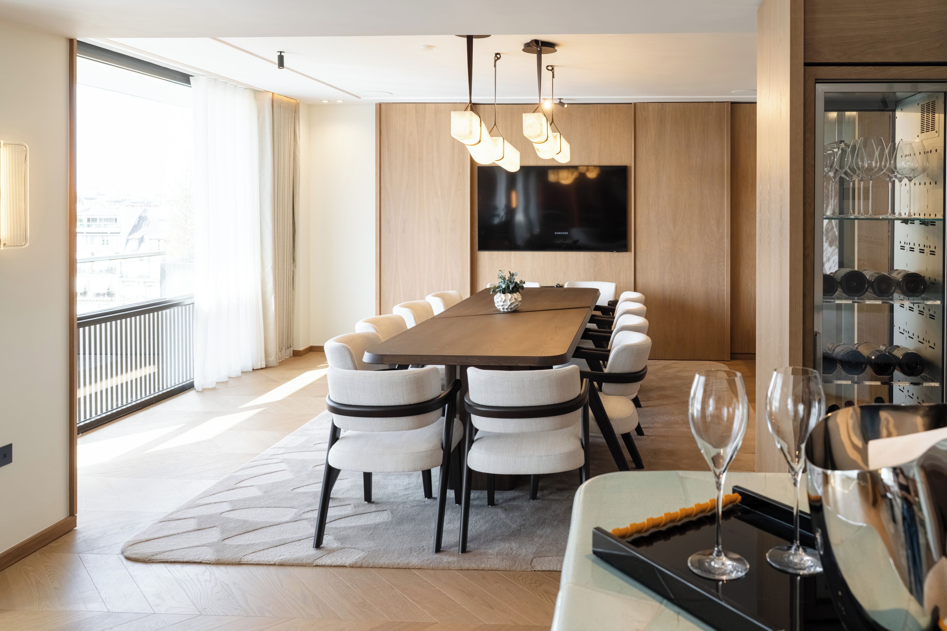 A modern dining area with a long wooden table, multiple chairs, a flat-screen TV mounted on the wall, and a wine rack with bottles.