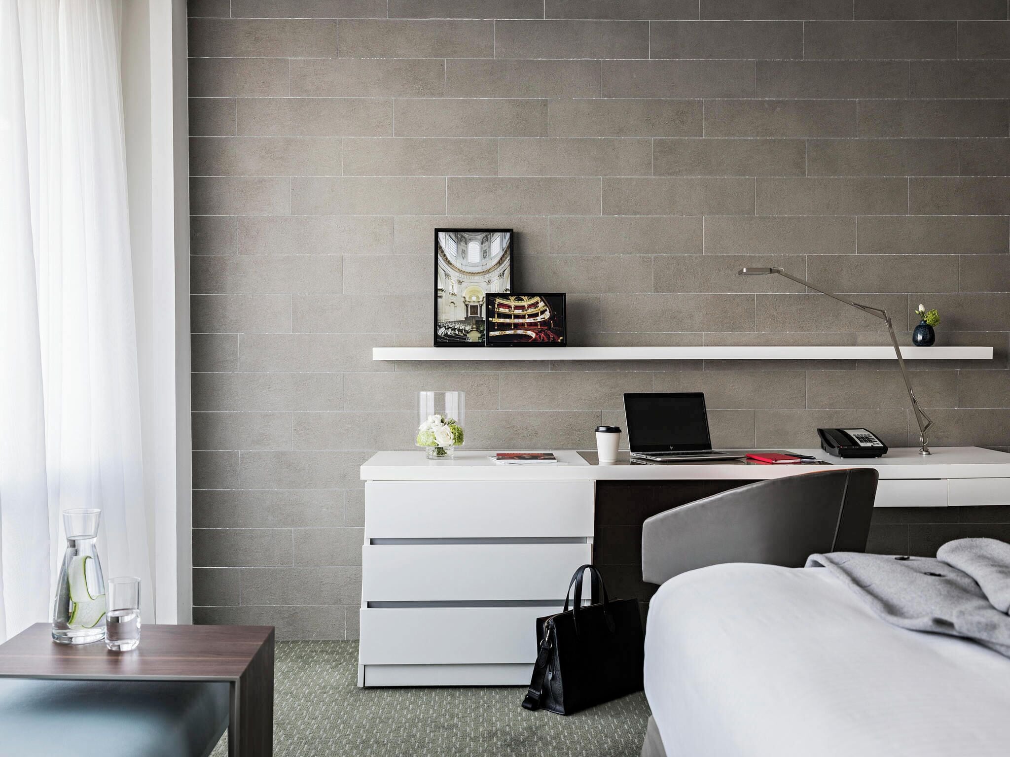 A modern hotel room with a grey brick wall, a white desk with a laptop, a phone, and a picture frame. There is a bed with a grey blanket and a black handbag on the floor.