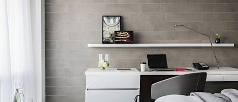 A modern hotel room with a grey brick wall, a white desk with a laptop, a phone, and a picture frame. There is a bed with a grey blanket and a black handbag on the floor.
