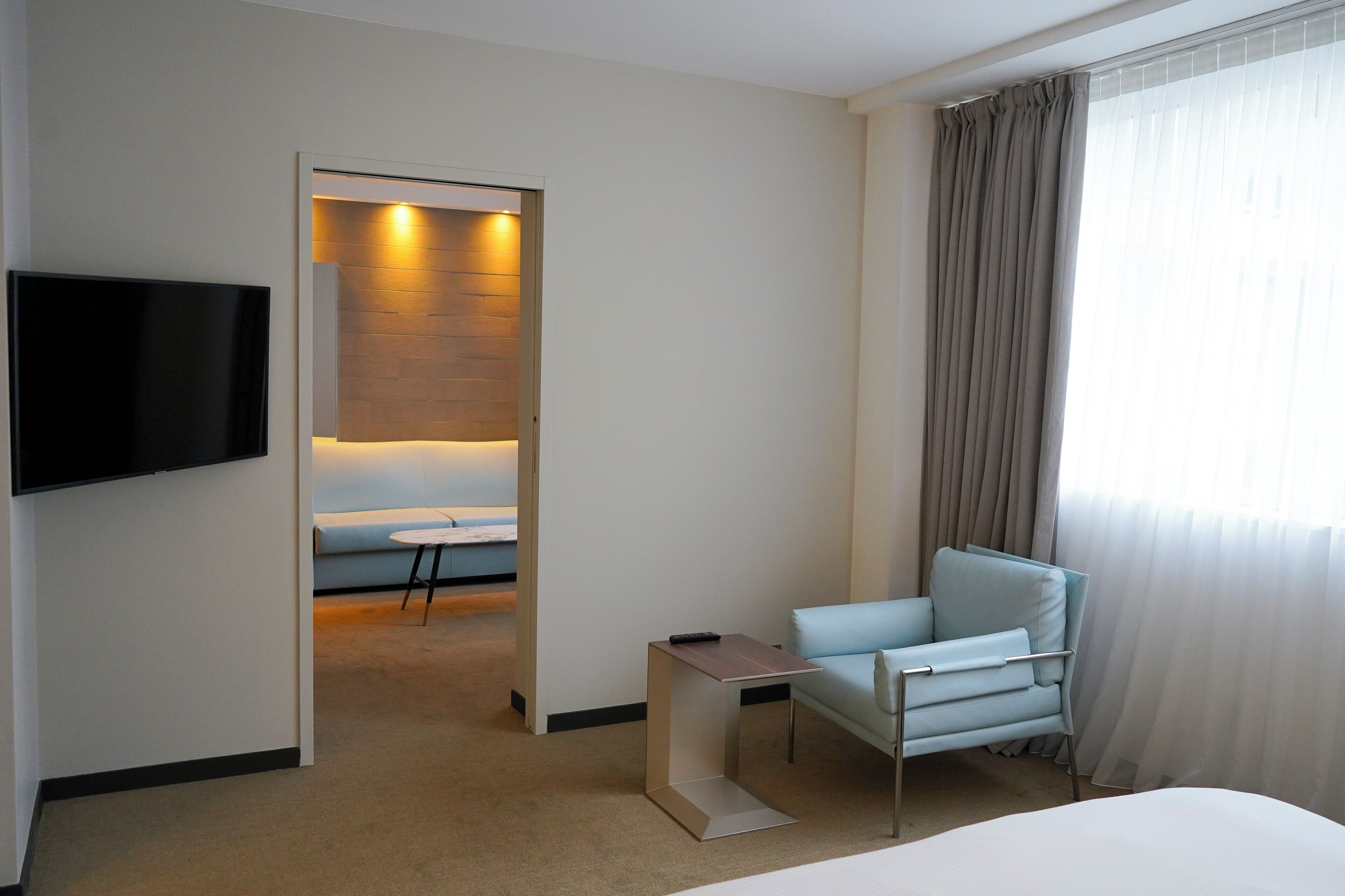 A modern hotel room with a flat-screen TV mounted on the wall, a light blue armchair, a small table, and a bed.