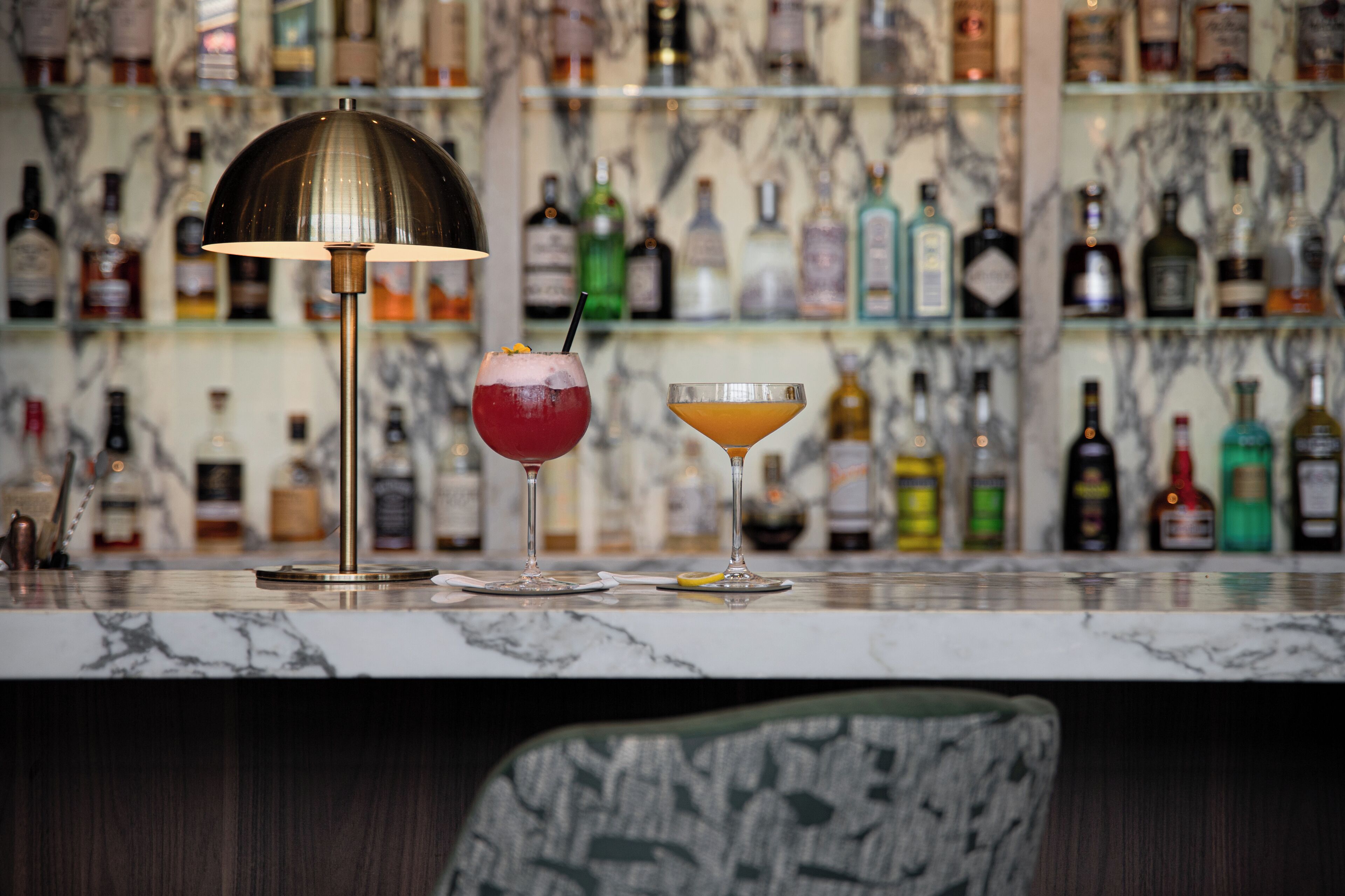A bar area with a marble countertop, two drinks, a lamp, and a shelf of liquor bottles.