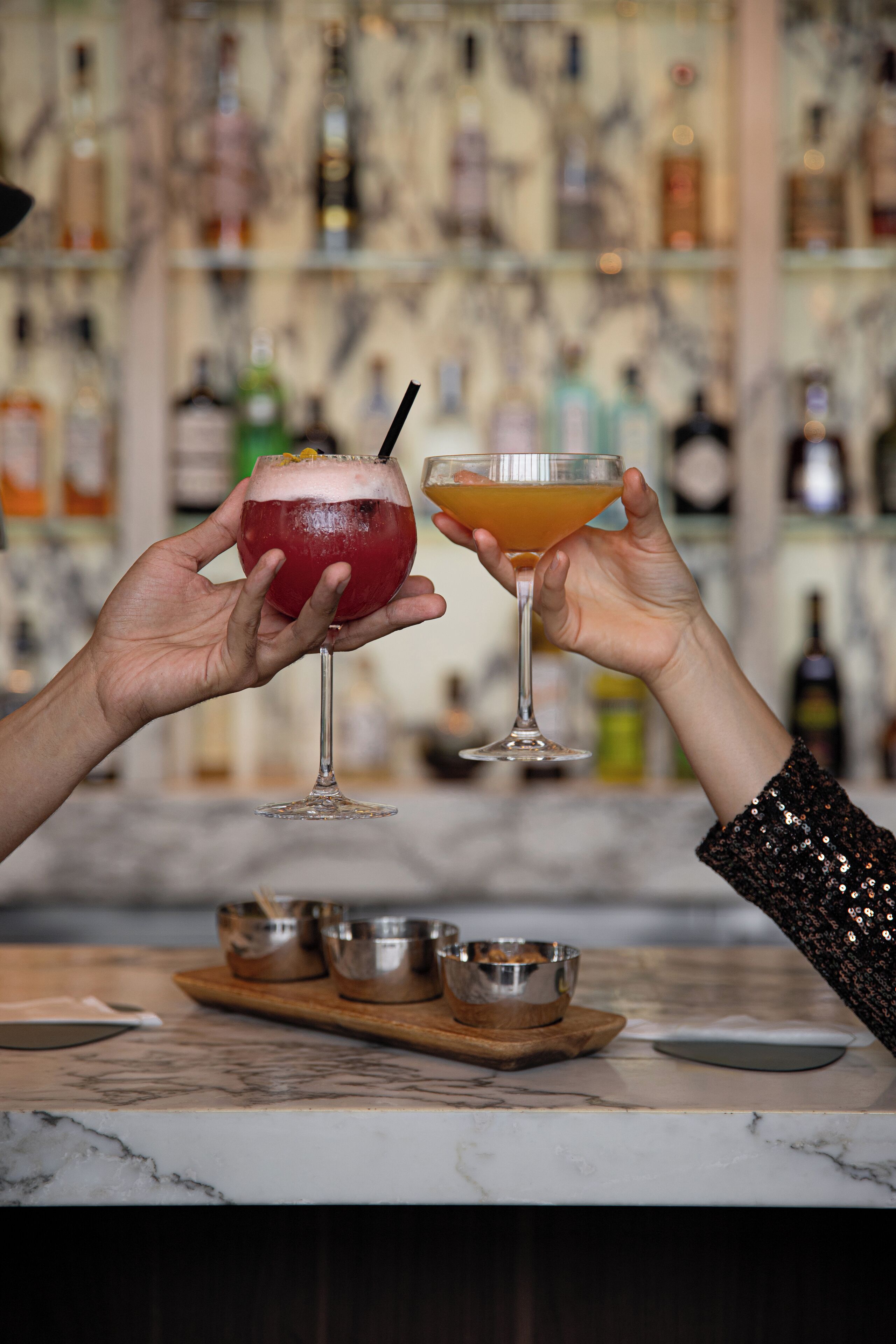 Duas mãos segurando copos de coquetel, um balcão de bar com tampo de mármore e uma estante com vários frascos ao fundo.