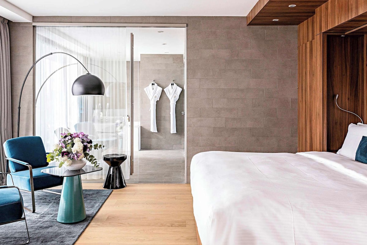 A modern hotel room with a large bed, a blue armchair, a small round table with a vase of flowers, and a large floor lamp.