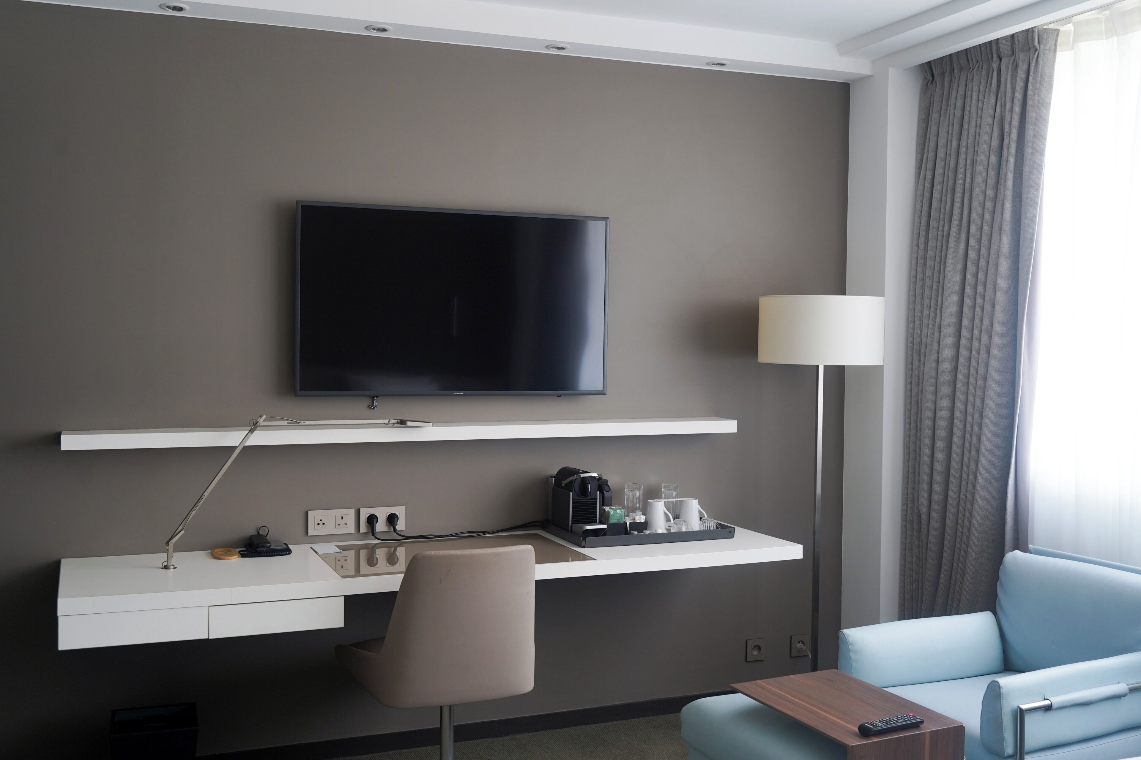 A hotel room with a flat-screen TV mounted on the wall, a desk with a lamp, a chair, a sofa, and a remote control on the sofa.