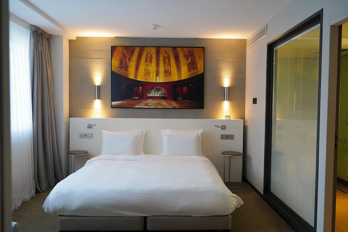 A hotel room with a large bed, two bedside tables, a wall-mounted TV, and a sliding glass door.