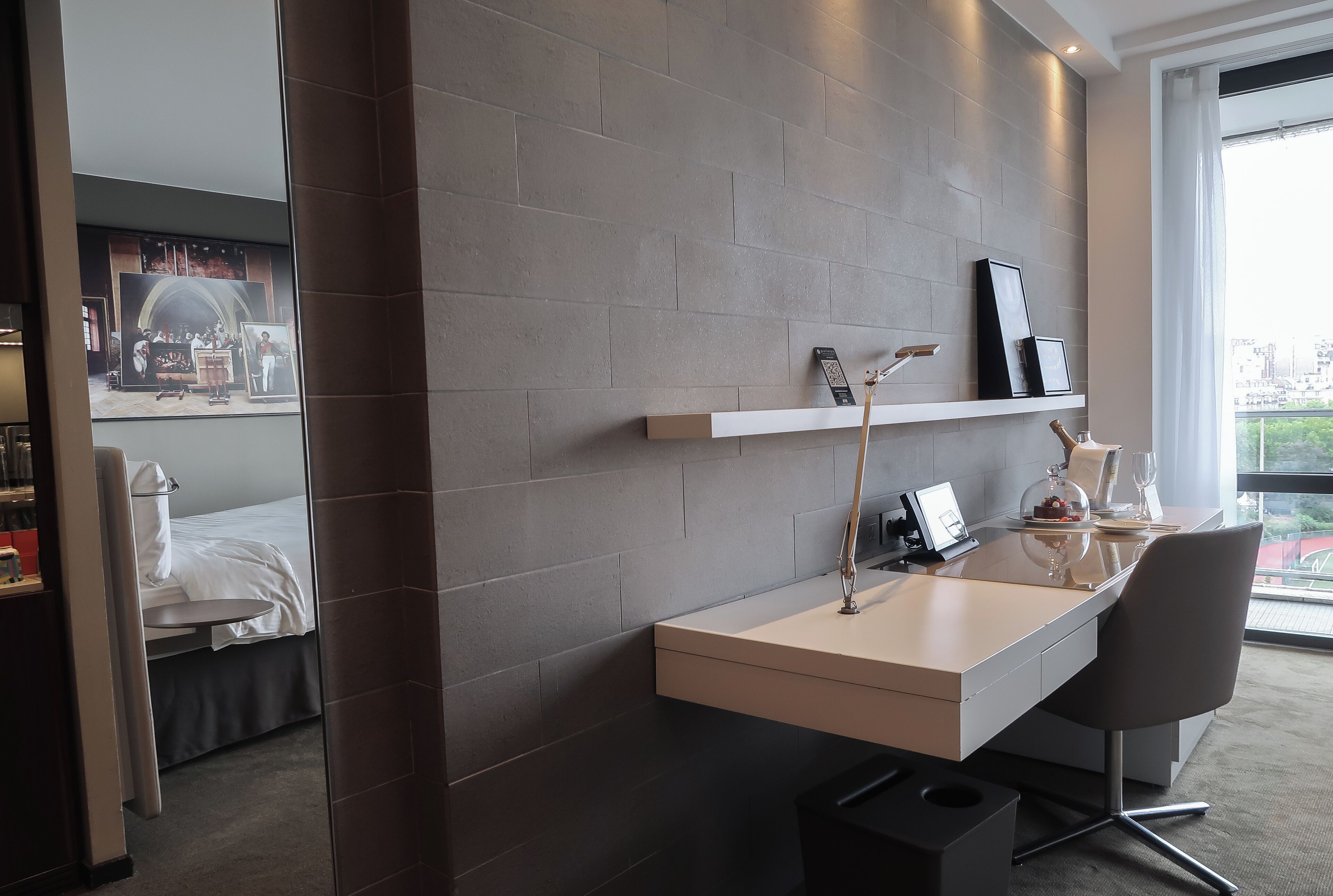 A modern hotel room with a minimalist design, featuring a built-in desk, a large window with a city view, and a cozy bed area.