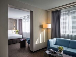 A hotel room with a bed, a blue sofa, a coffee table with a magazine and a vase of flowers, a lamp, and a view of another room.