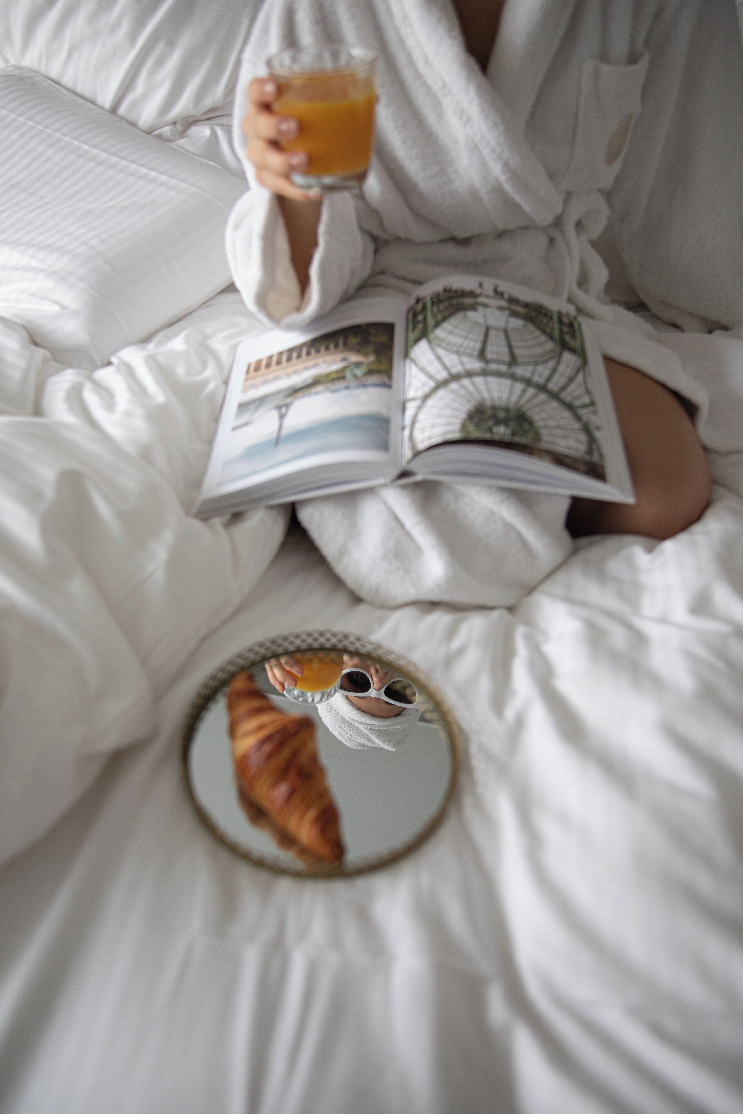Eine Person in einem weißen Bademantel sitzt auf einem Bett mit einem Buch und einem Glas Orangensaft.