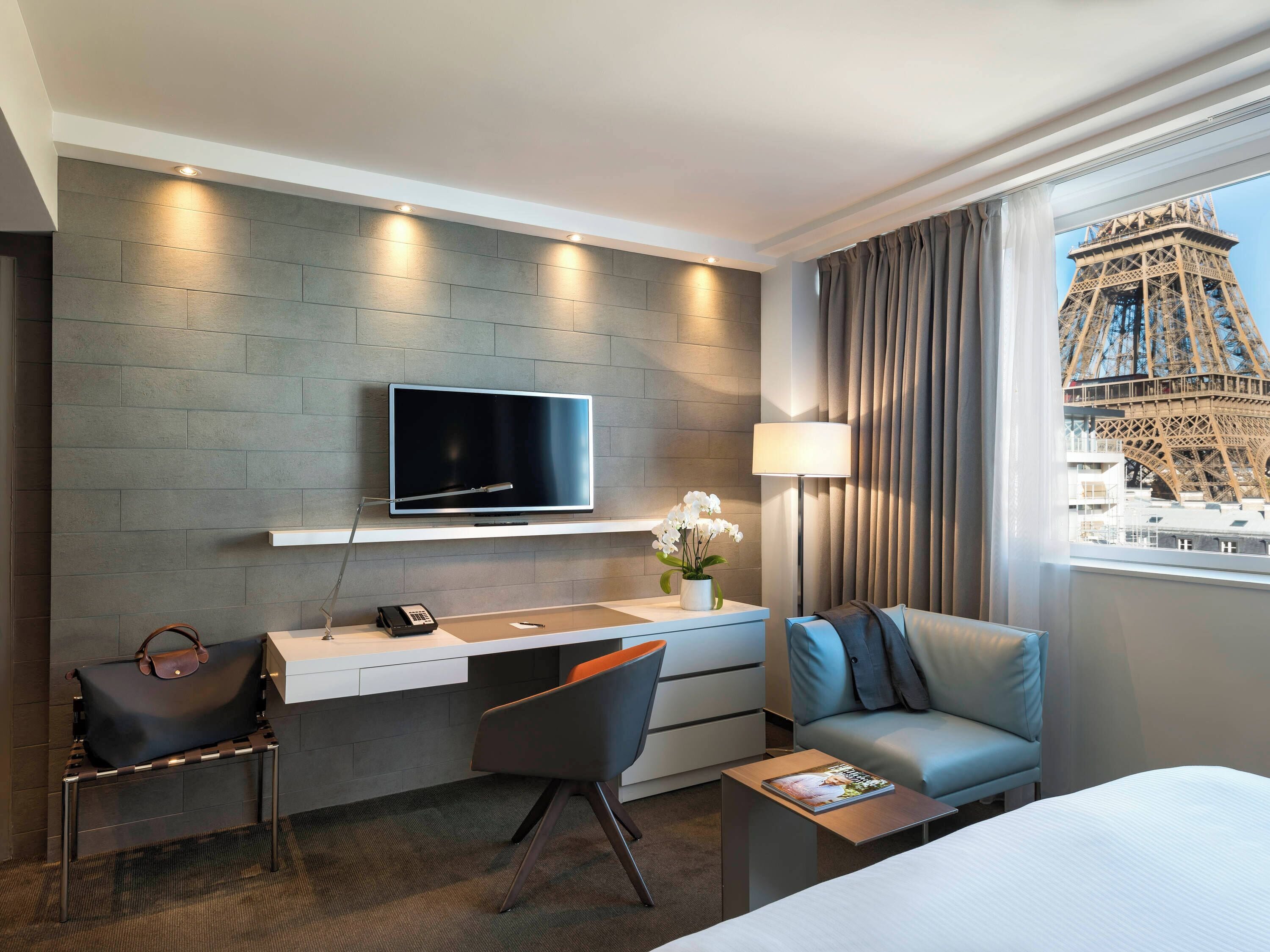 A modern hotel room with a flat-screen TV mounted on a grey brick wall, a desk with a telephone, a chair, a sofa, and a view of the Eiffel Tower through a window.