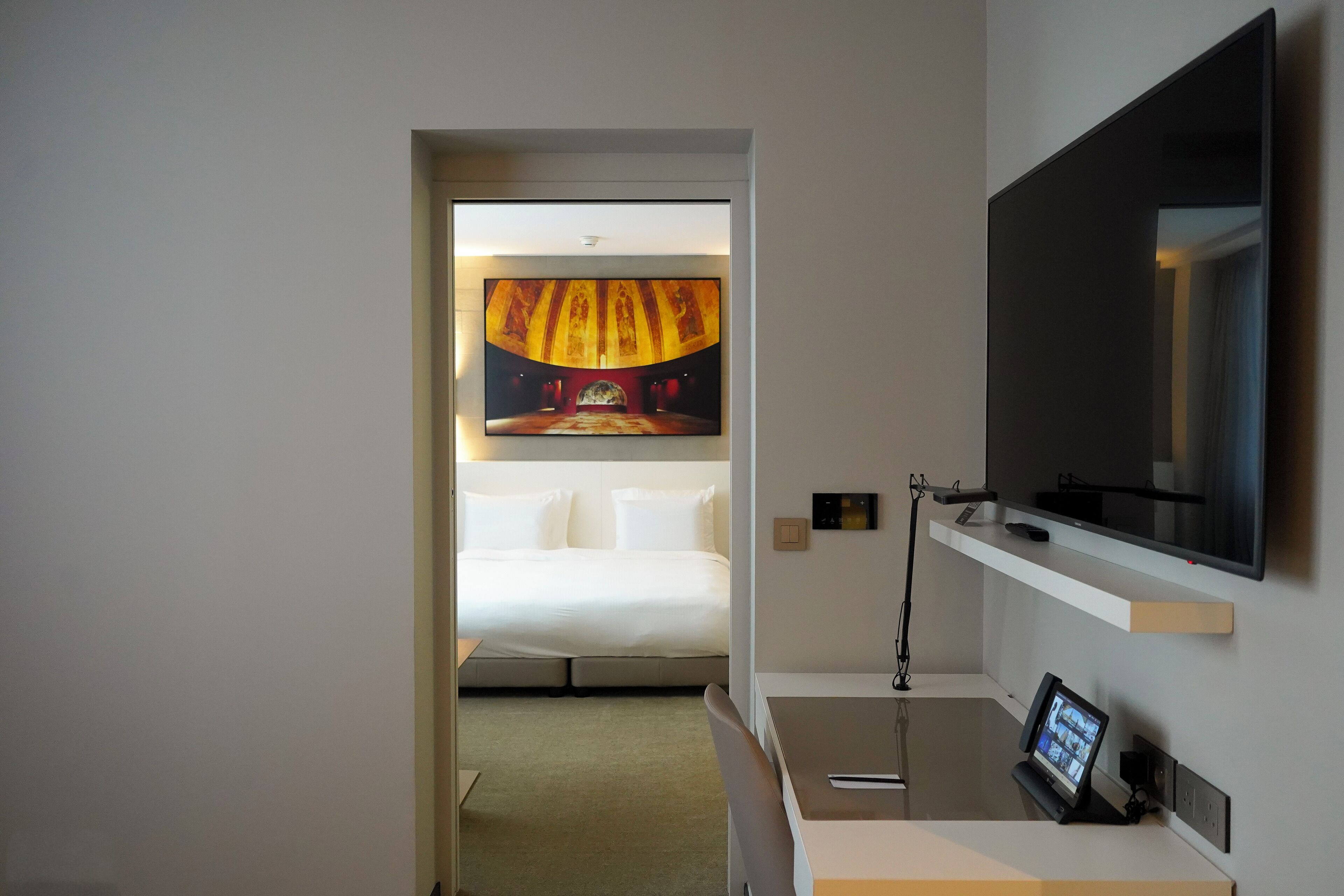 A hotel room with a bed, a TV mounted on the wall, a desk with a computer, and a painting of a theater on the wall.
