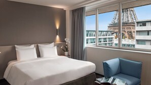 Camera d'albergo con un letto grande, una poltrona blu, un comodino con una lampada e vista sulla Torre Eiffel.