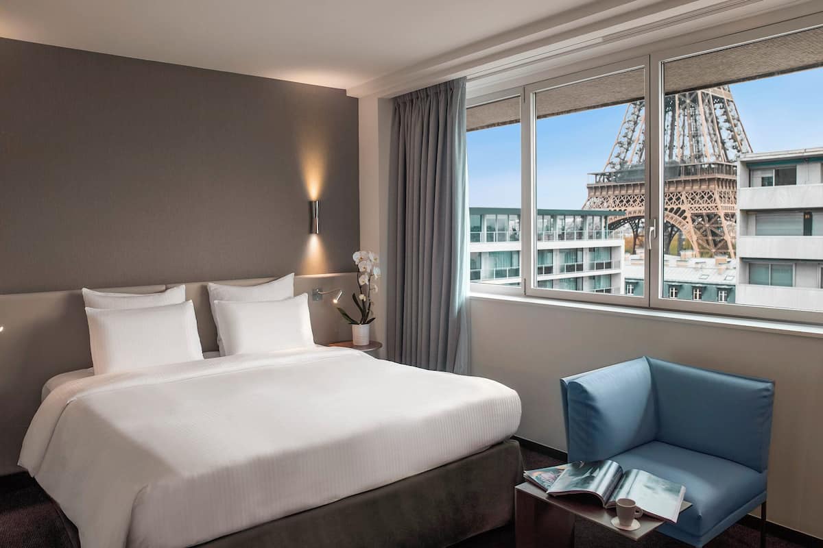 A hotel room with a large bed, a blue armchair, a nightstand with a lamp, and a view of the Eiffel Tower.
