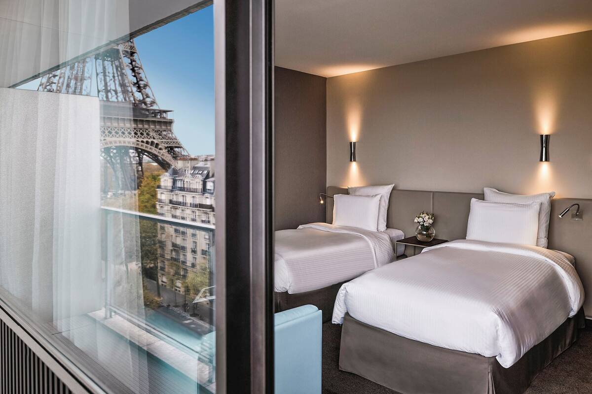 A hotel room with two beds, a view of the Eiffel Tower, and modern wall sconces.