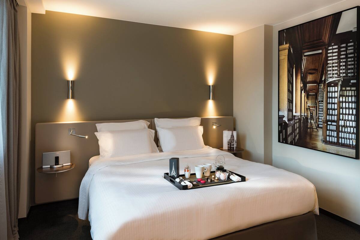 A hotel room with a large bed, two bedside tables, a wall-mounted light fixture, and a framed picture of a library.