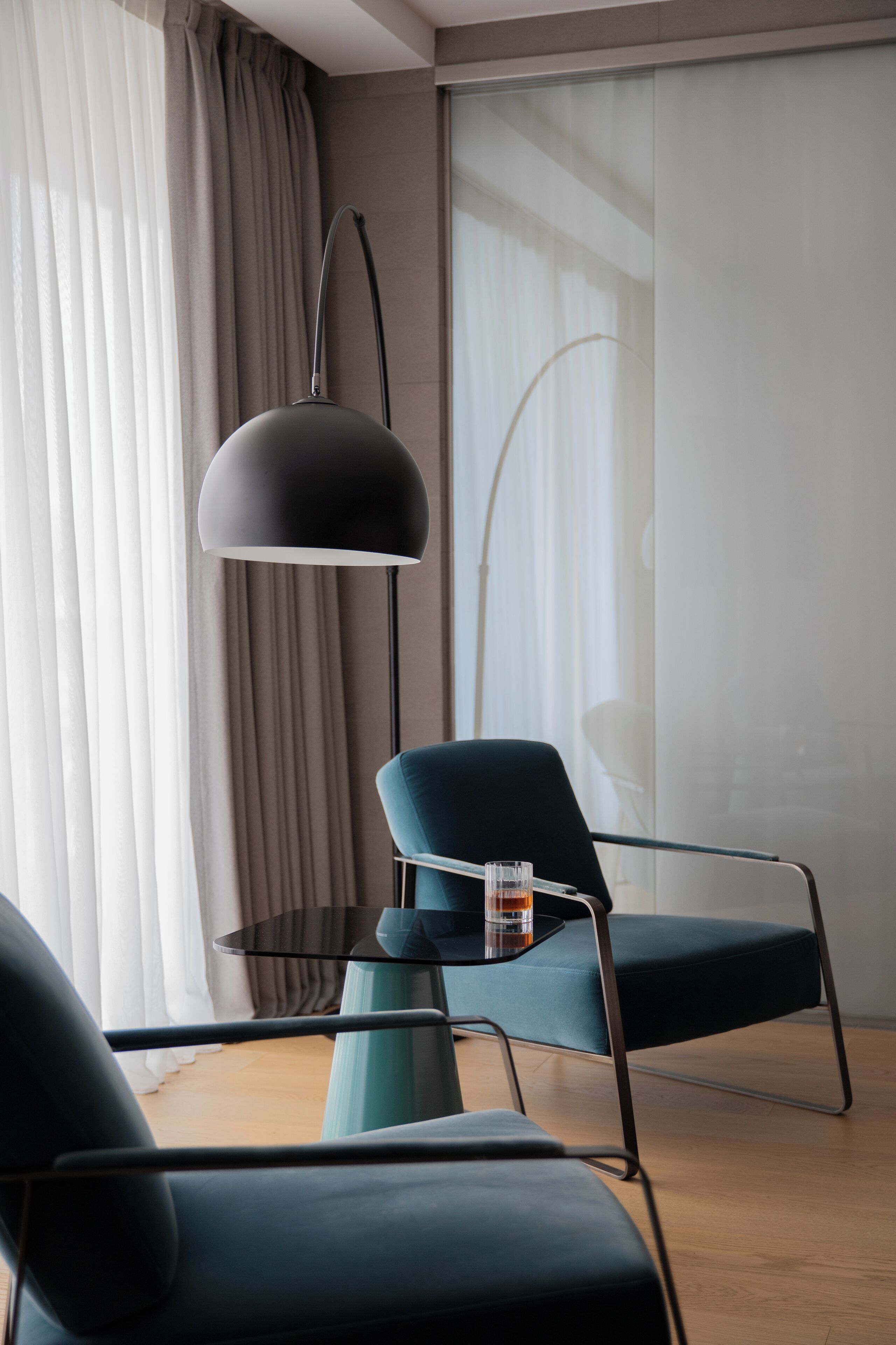 Een moderne kamer met een ronde glazen tafel, twee blauwgroene stoelen, een zwarte vloerlamp en een drankje.