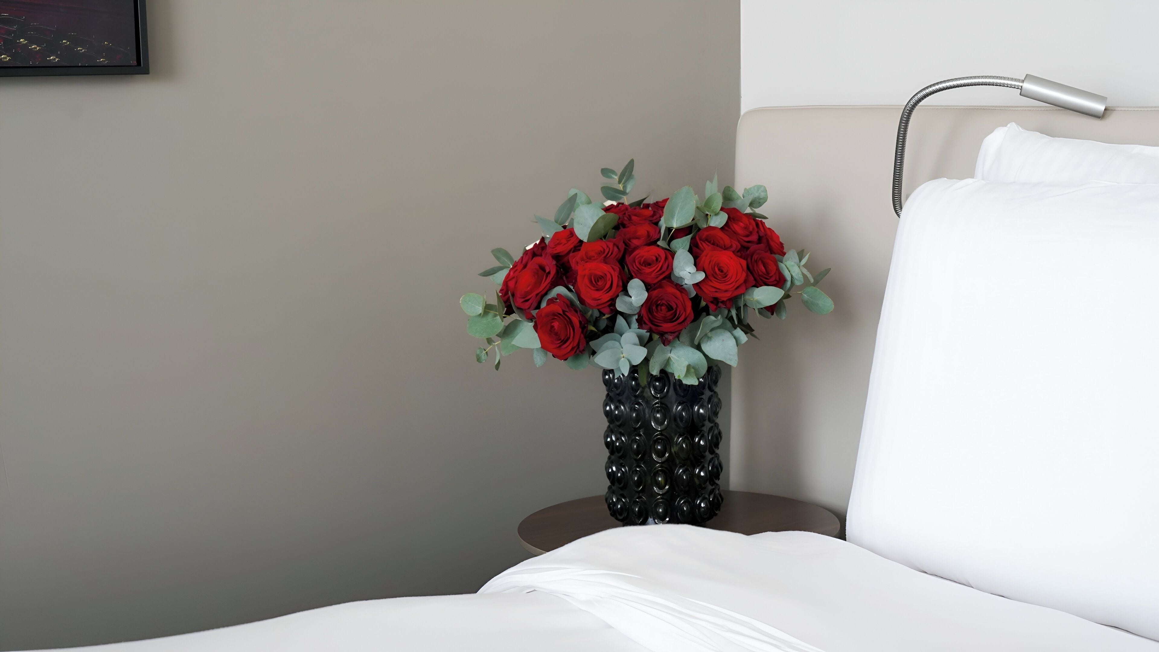 Un lit avec du linge de lit blanc, un vase contenant des roses rouges et des feuilles vertes, et un luminaire fixé au mur.