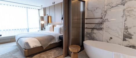 A modern bedroom with a large bed, a wooden stool, and a freestanding bathtub.
