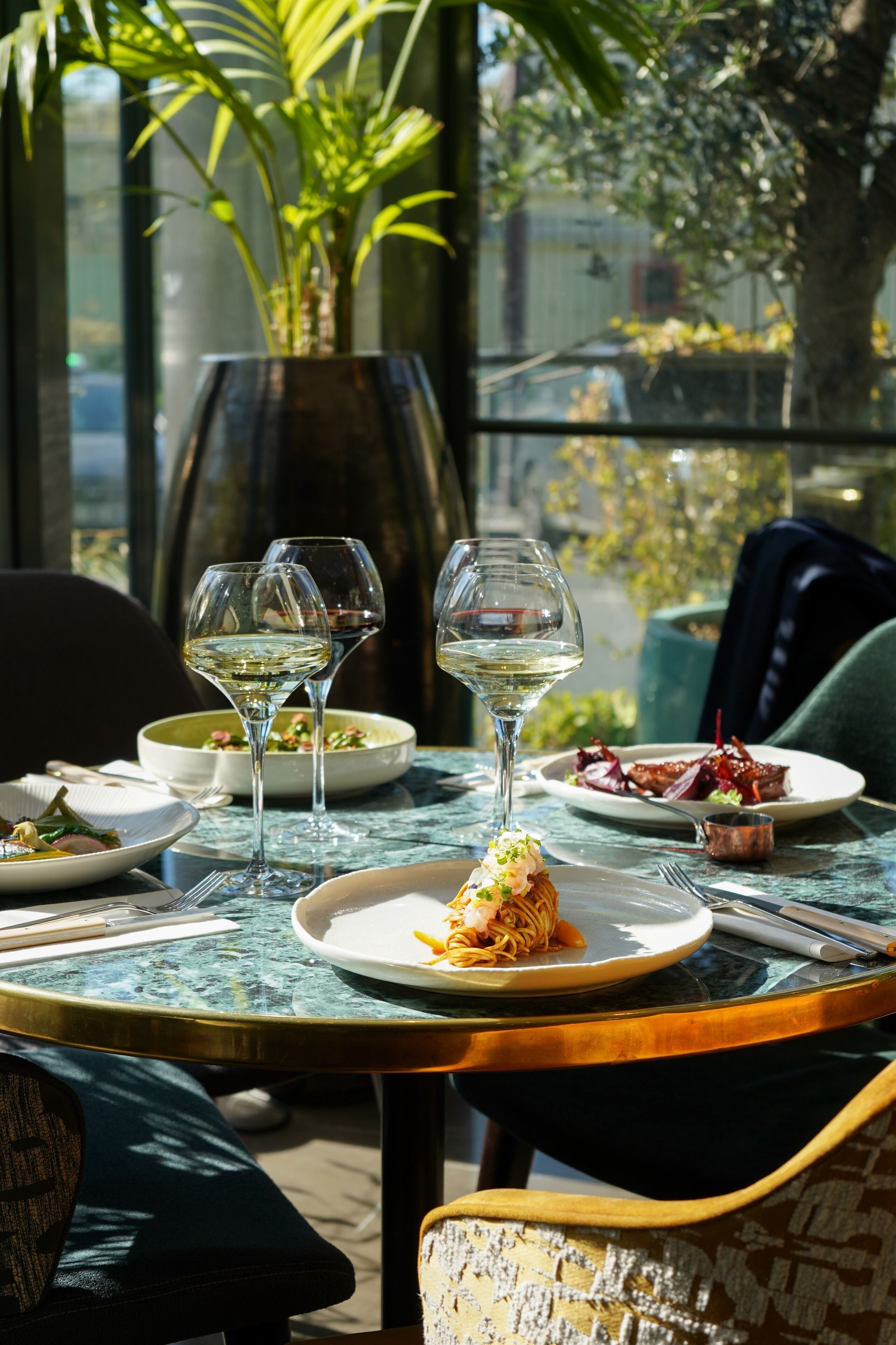 A dining table with plates of food and wine glasses, set in a room with a view of greenery outside.