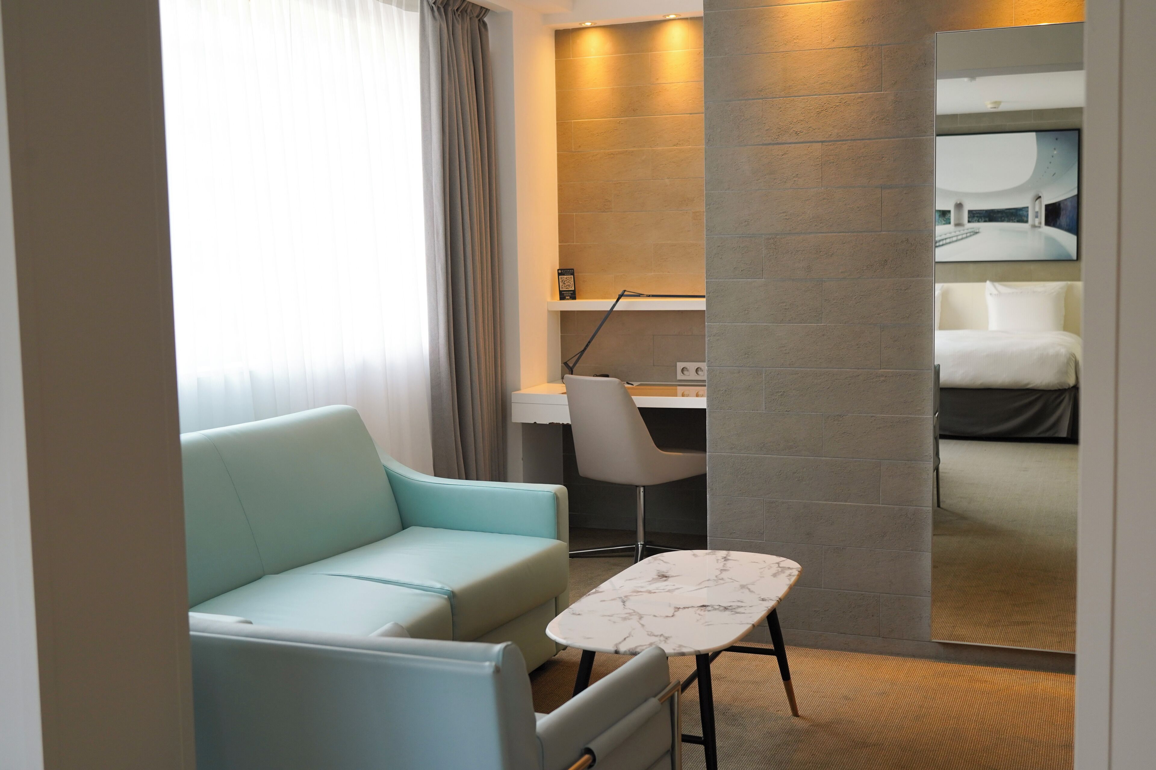 Une chambre d’hôtel moderne avec un canapé bleu clair, une petite table en marbre, un bureau avec une chaise et un grand miroir qui reflète un lit.