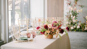 A table set for a meal with a floral centerpiece, clear chairs, and a view of the outdoors.