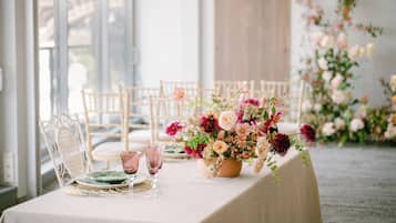 Une table dressée pour un repas, agrémentée d’un centre de table floral, de chaises transparentes et offrant une vue sur l’extérieur.