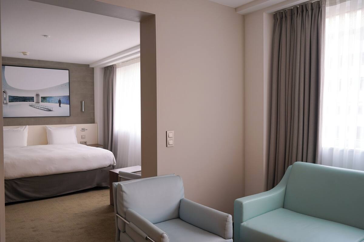 A hotel room with a large bed, a desk, two armchairs, a flat-screen TV mounted on the wall, and a window with curtains.