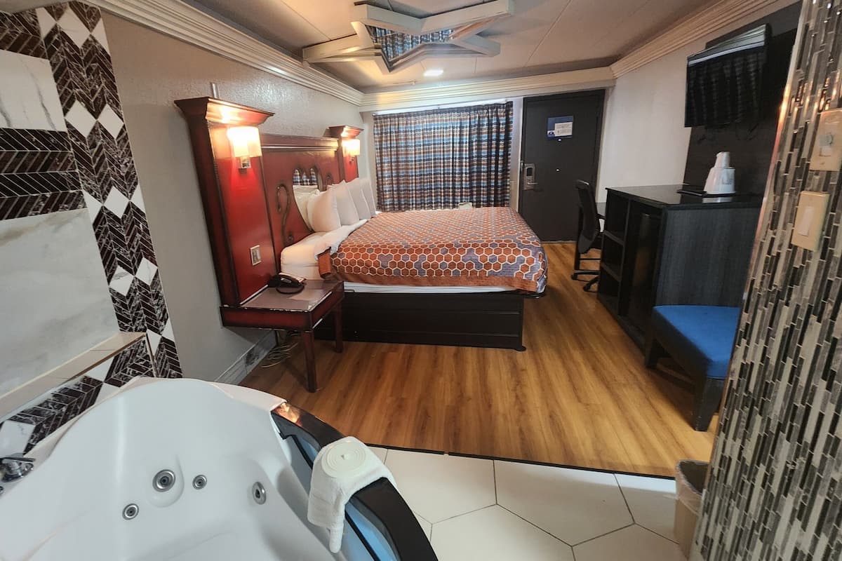 A compact hotel room with a bed, a desk, a chair, a vanity with a mirror, and a small bathroom with a bathtub.