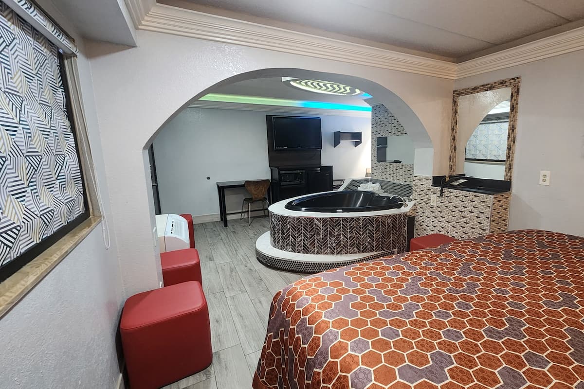 A hotel room with a large bed, a sitting area with a red chair, a small desk, a bathtub, and a television.