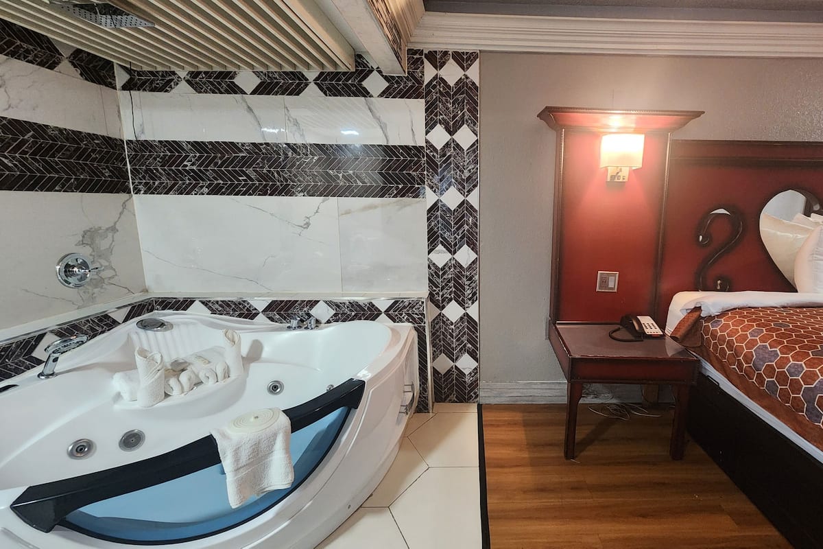 A hotel room with a large bathtub, a bed with a patterned bedspread, a bedside table with a lamp, and a wall with a geometric tile pattern.