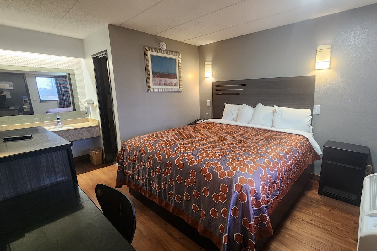 A hotel room with a bed, a desk, a chair, a mirror, and a wall-mounted heater.