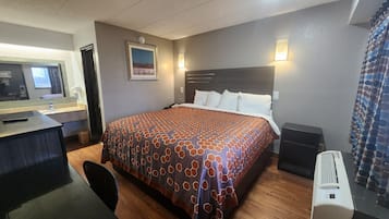 A hotel room with a bed, a desk, a chair, a mirror, and a wall-mounted heater.