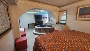 Une chambre d’hôtel avec un grand lit, un coin salon avec un fauteuil rouge, une petite table, un téléviseur et un jacuzzi.