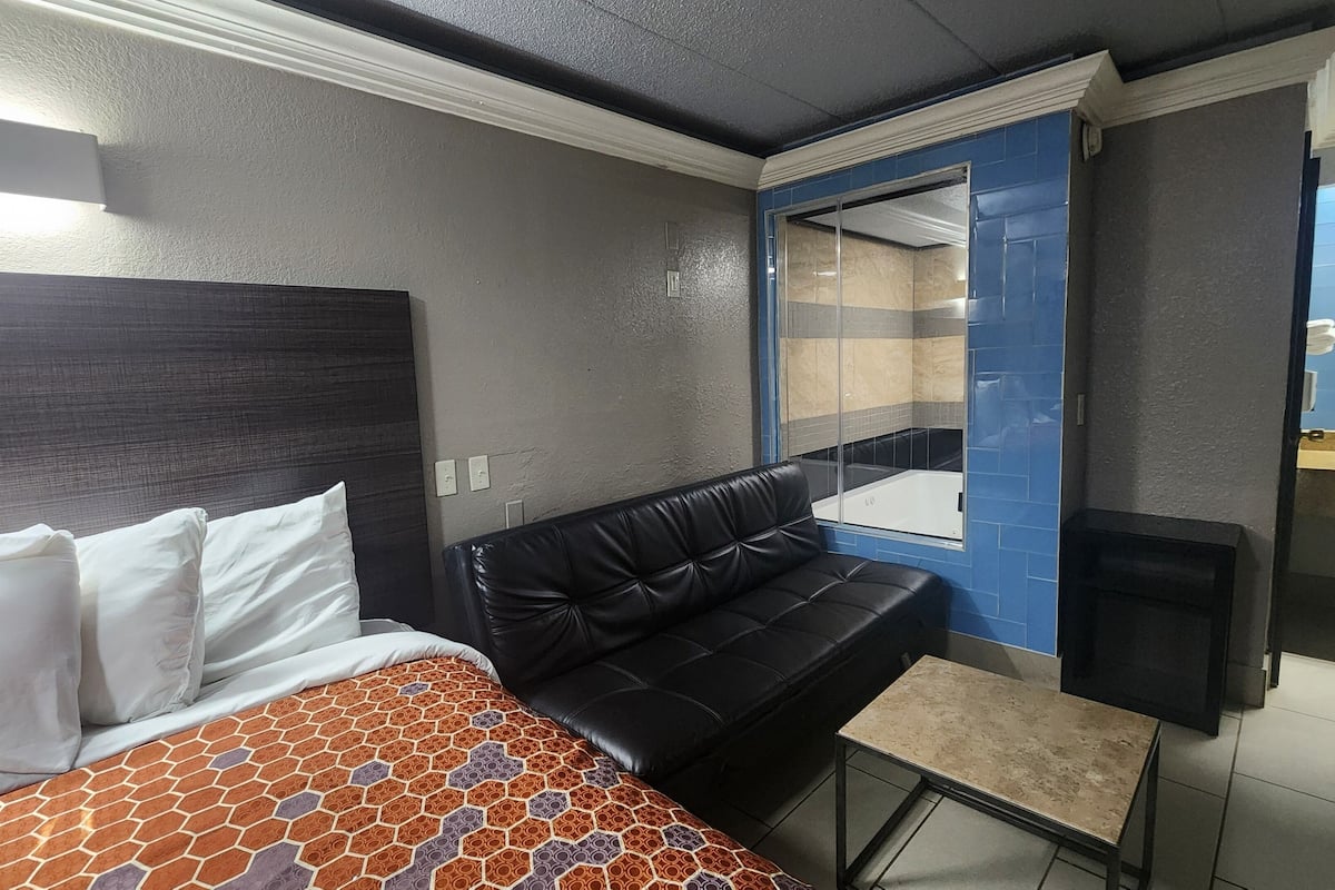 A hotel room with a bed, a black leather sofa, a small table, and a bathroom visible through a glass door.