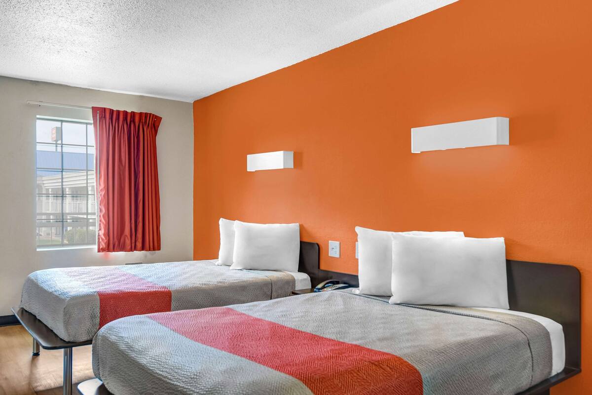 Habitación de hotel con dos camas, paredes anaranjadas y una ventana con cortinas rojas.