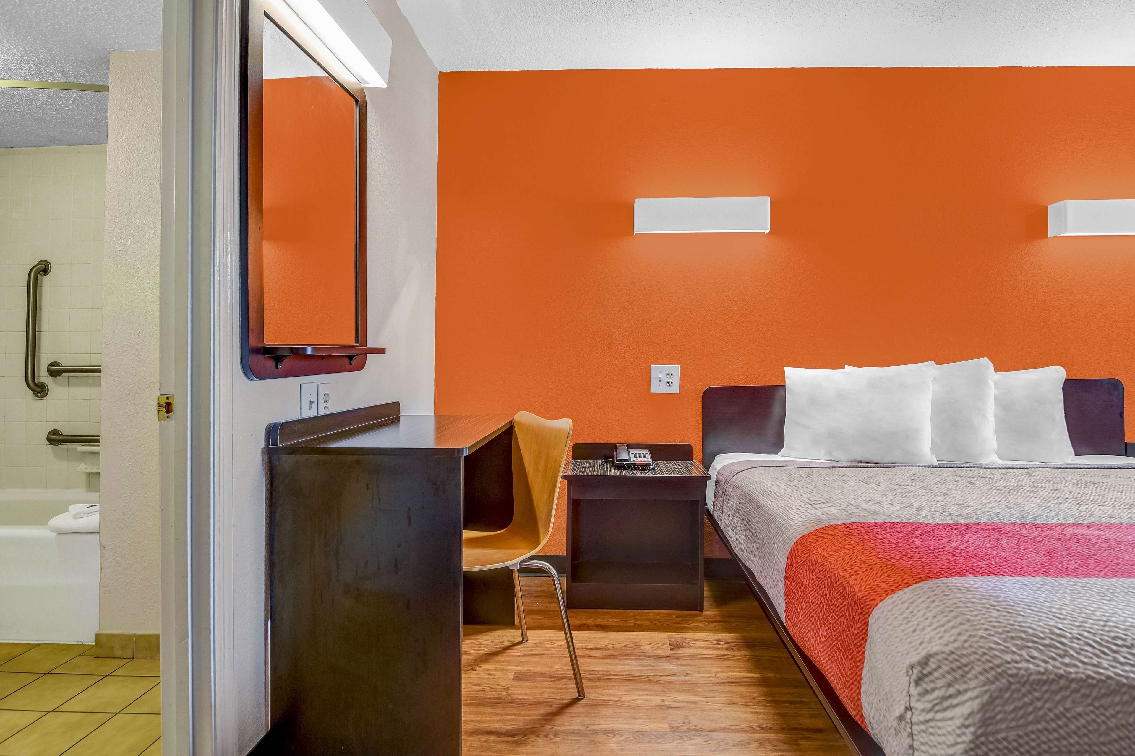 A hotel room with a bed, a desk, a chair, a small table, a mirror, and a bathroom with a shower and a toilet.