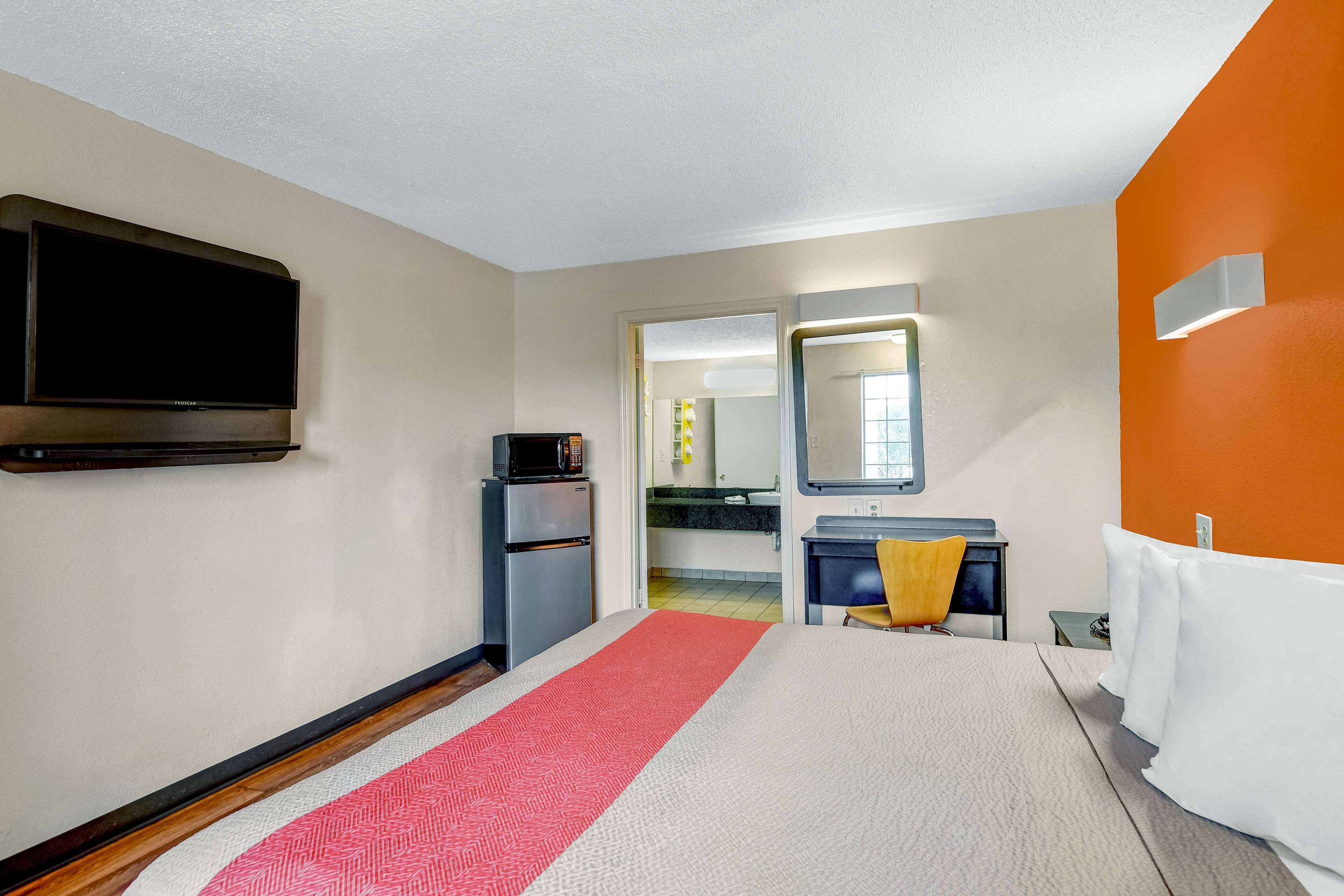 A hotel room with a bed, a TV, a refrigerator, a microwave, a sink, and an orange accent wall.
