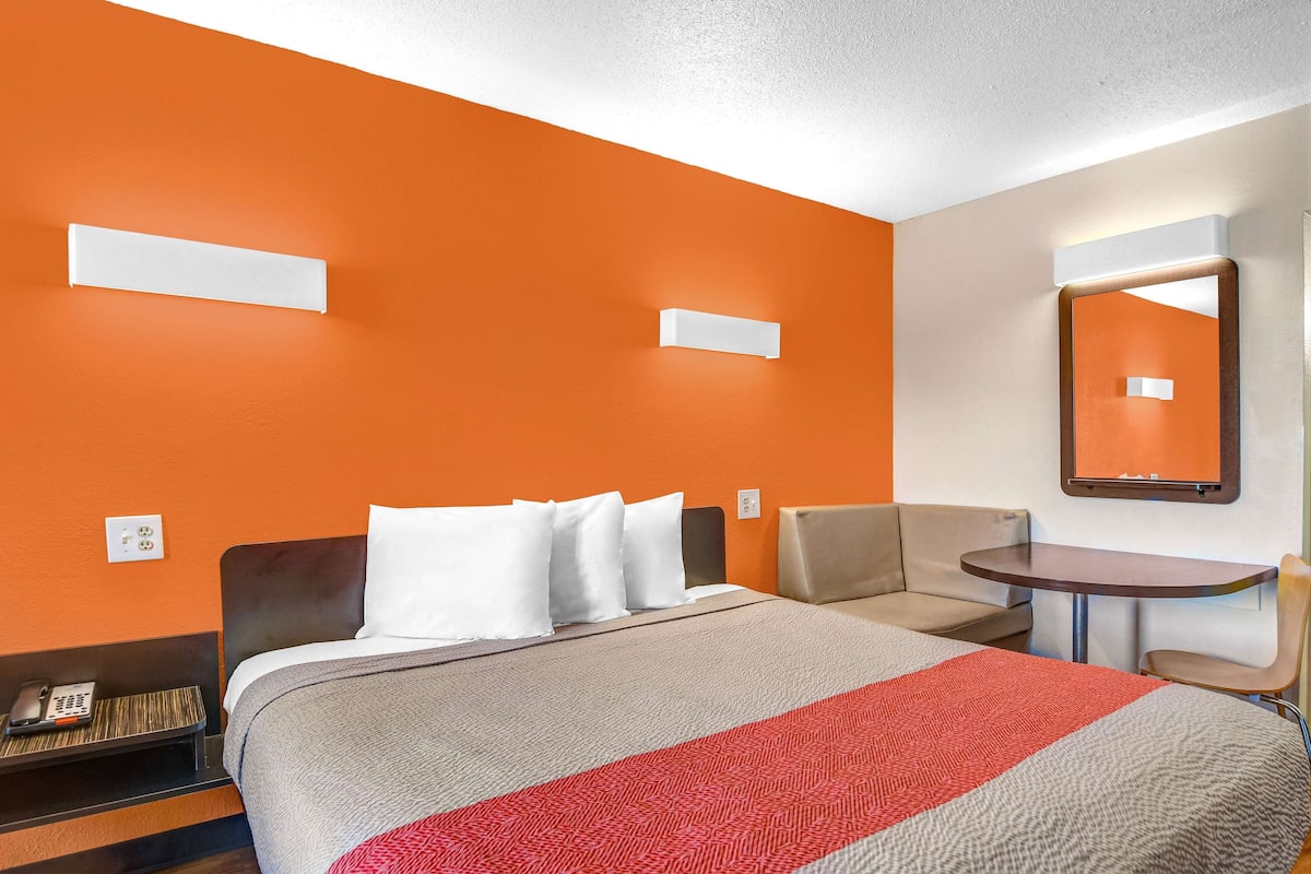 Una habitación de hotel con una pared naranja, una cama con ropa de cama blanca y gris, una mesita, una silla y un espejo.