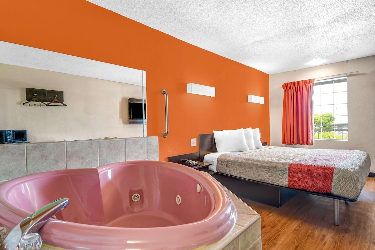 Una habitación de hotel con una bañera rosa grande, una cama con manta gris y roja, y una ventana con cortinas naranjas.