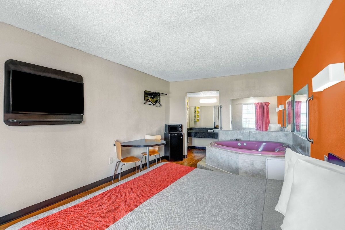 Habitación de hotel con cama, escritorio, silla, televisor, microondas, baño con bañera y ventana con cortinas.