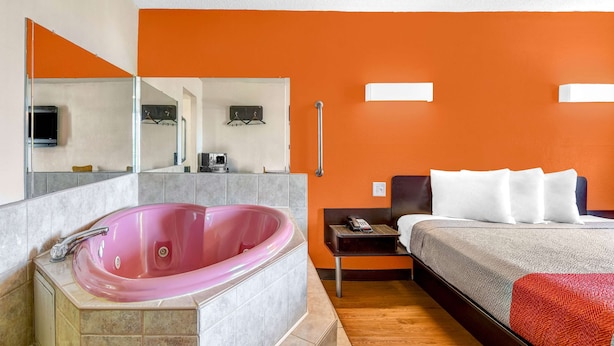 A modern hotel room with a large pink jacuzzi, a bed with white bedding, and an orange accent wall. - Motel 6 Calhoun, GA (Calhoun)
