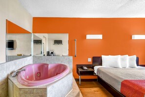 A modern hotel room with a large pink jacuzzi, a bed with white bedding, and an orange accent wall.
