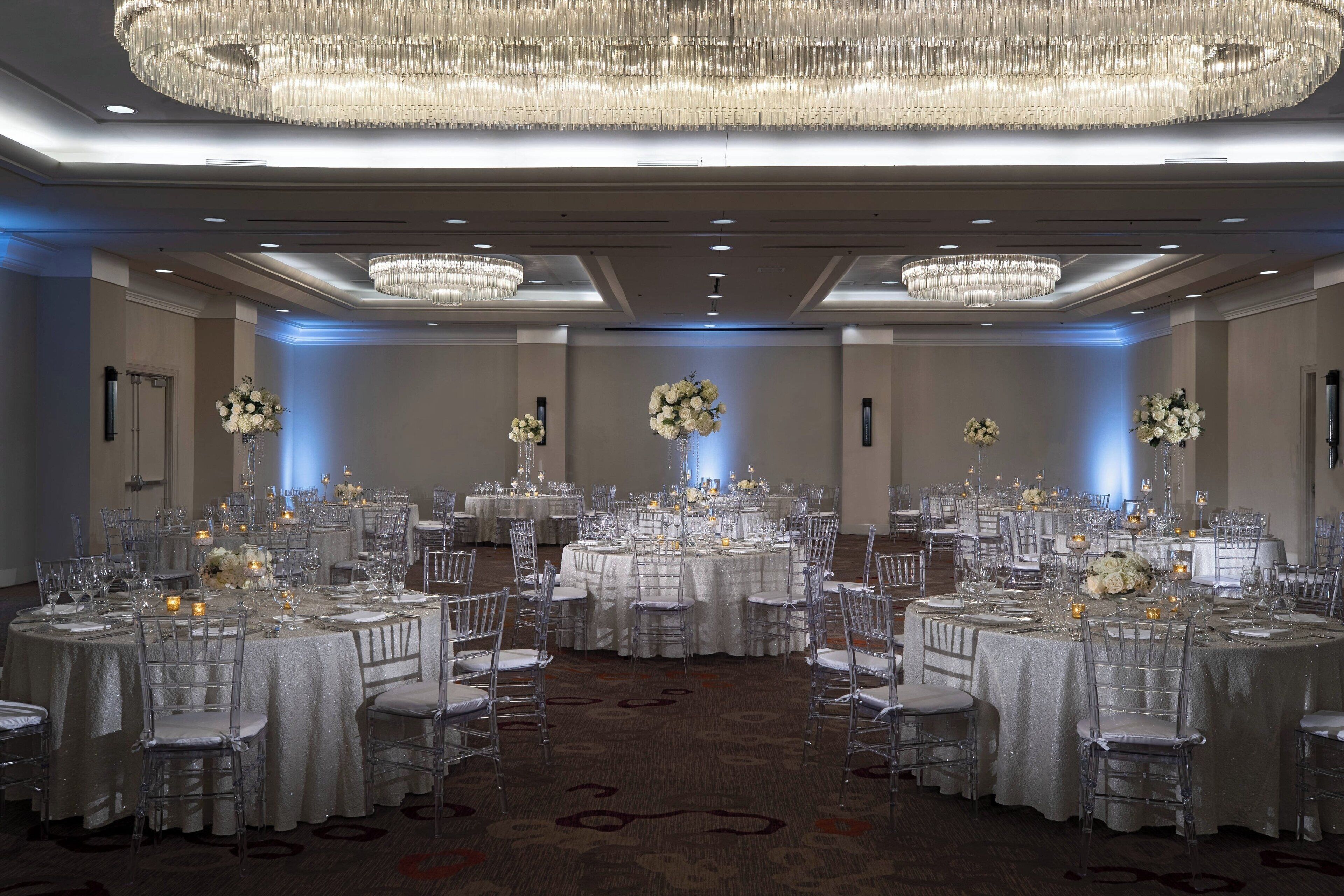 A banquet hall with round tables, clear chairs, and chandeliers.