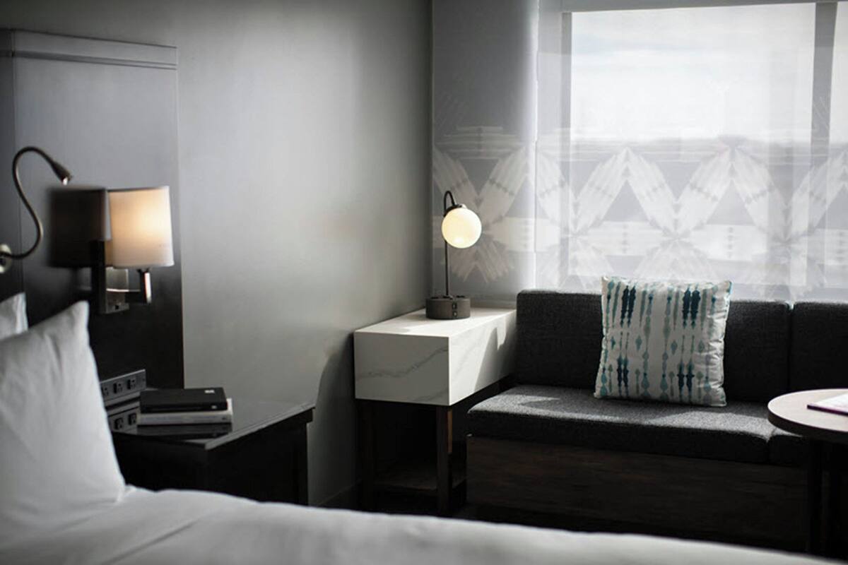 A modern hotel room with a bed, a bedside table with a lamp, a small table with a lamp, a sofa with a patterned pillow, and a window with a geometric view.