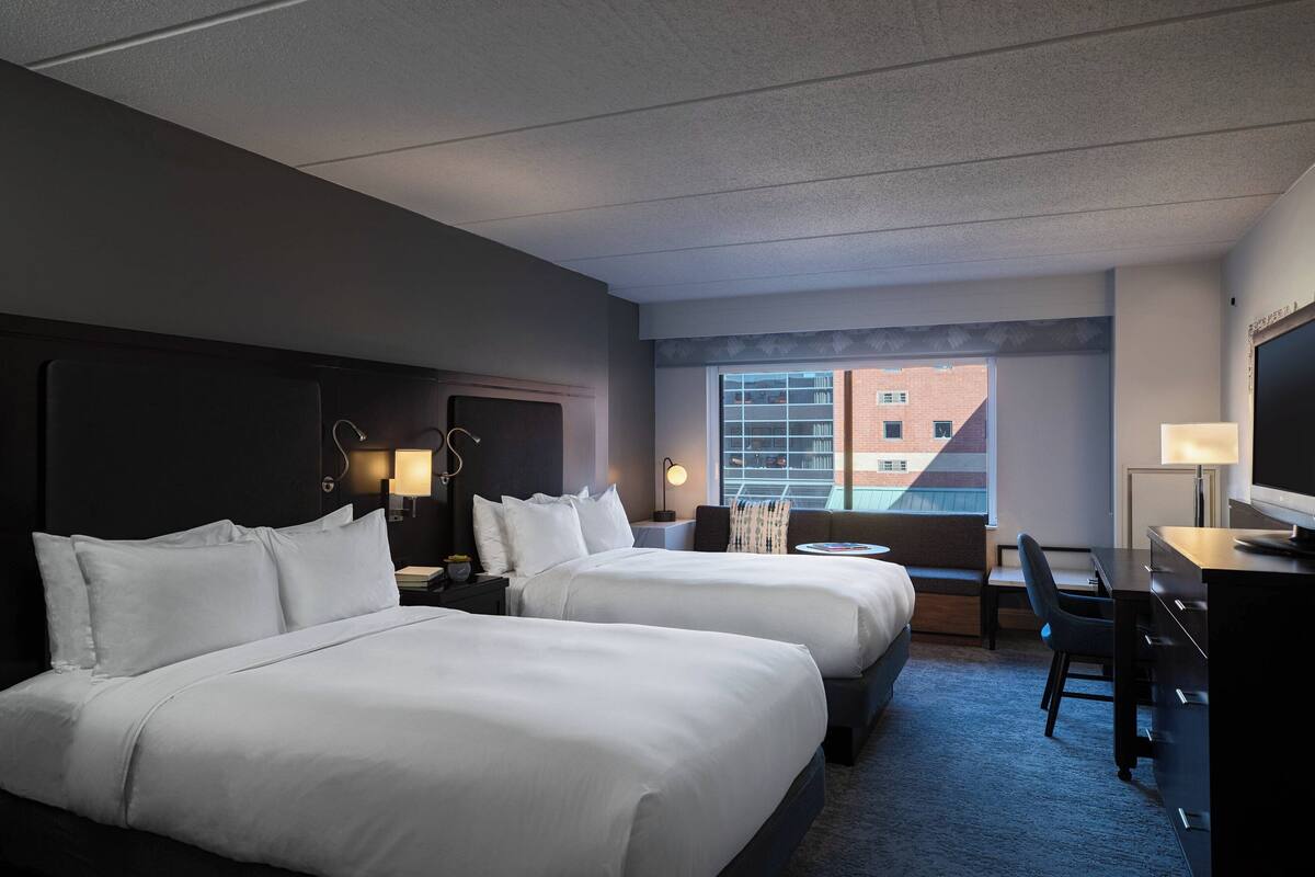 A hotel room with two beds, a desk, a chair, a TV, and a window with a view of a building.