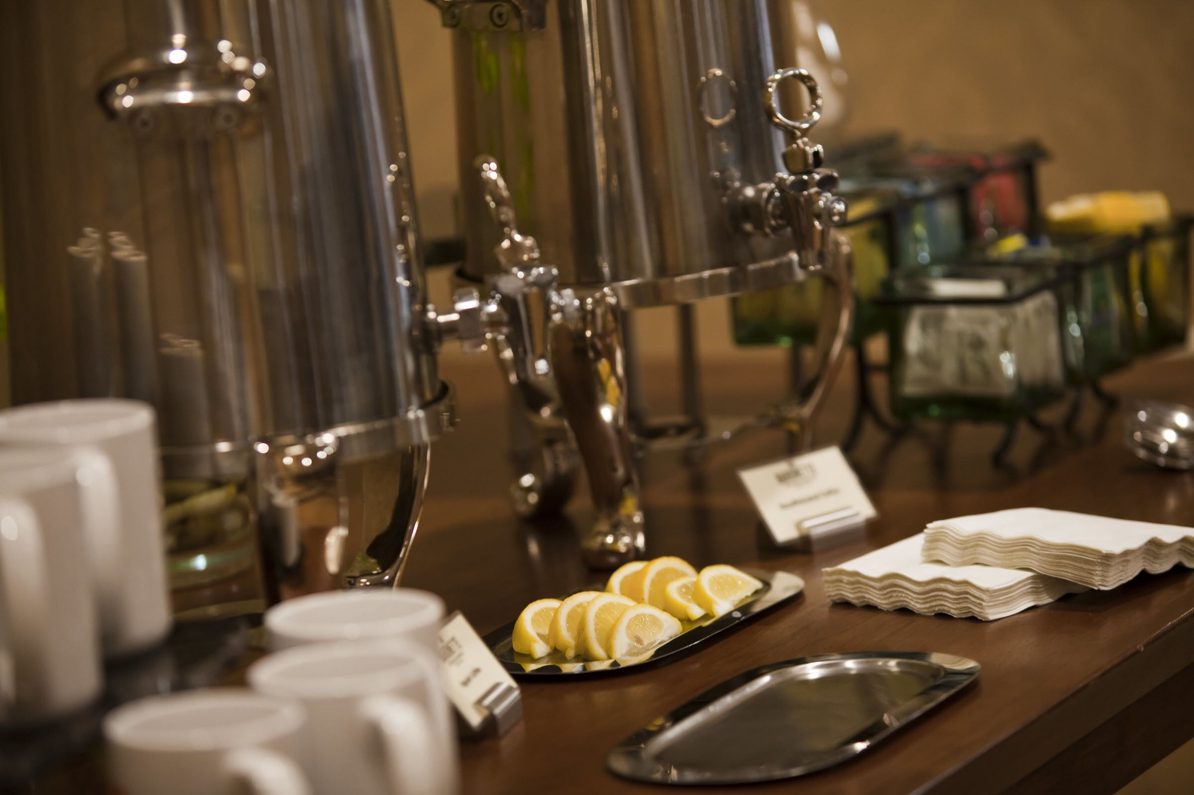 Buffet med metaldispenser, fad med citronskiver, stabel af servietter og flere hvide kopper.