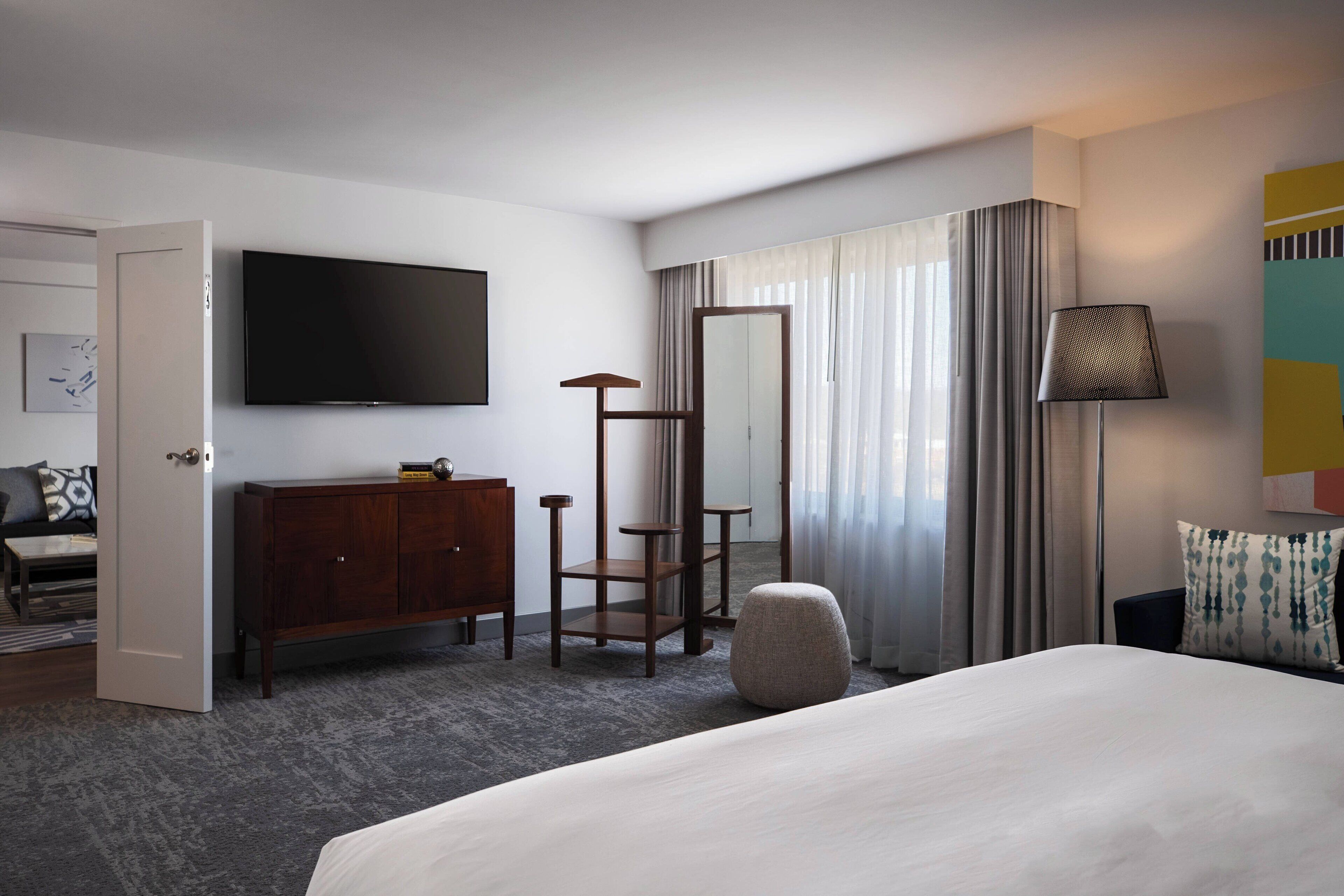 Une chambre d’hôtel avec un lit, une télévision, une commode, un miroir, une lampe et une fenêtre avec des rideaux.