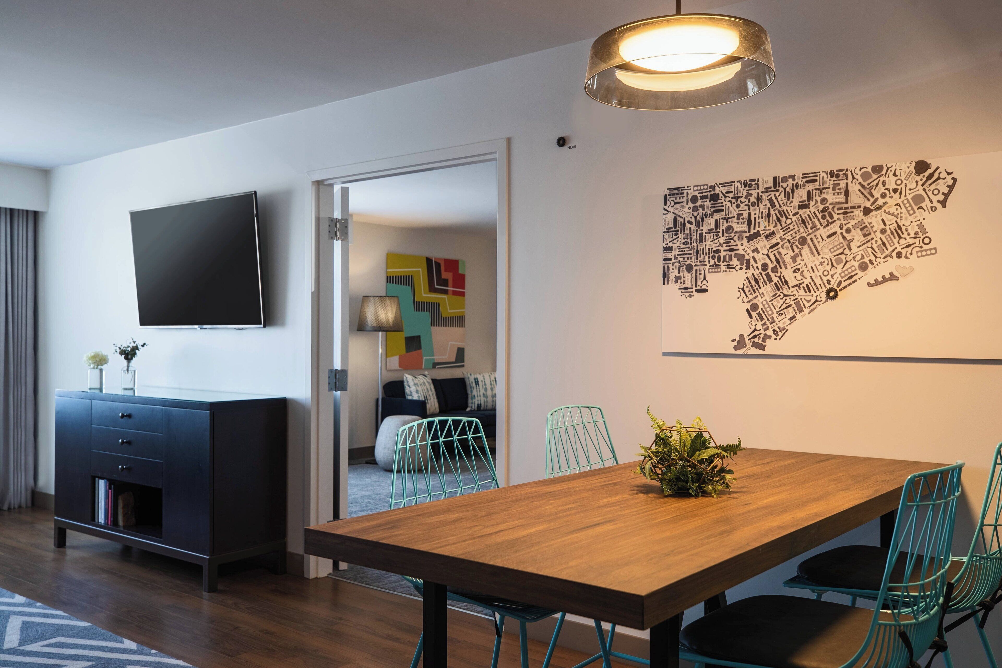 A modern dining area with a wooden table and teal chairs, a flat-screen TV on the wall, and a cityscape artwork.