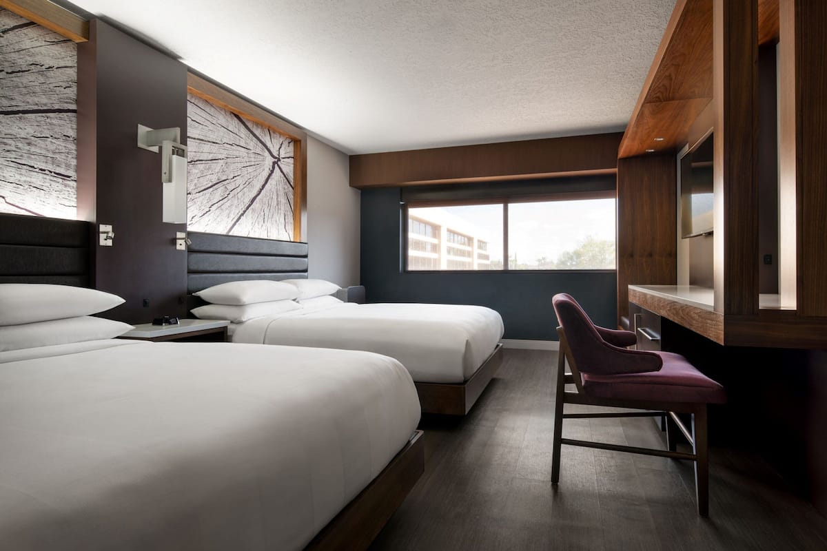 Ein Hotelzimmer mit zwei Betten, einem Schreibtisch, einem Sessel und einem großen Fenster mit Blick auf Gebäude.